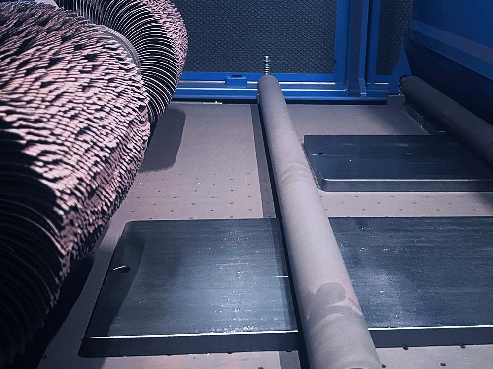 Innenansicht einer Maschine mit rotierenden Schleifbürsten und Metallplatten auf einem Förderband.
