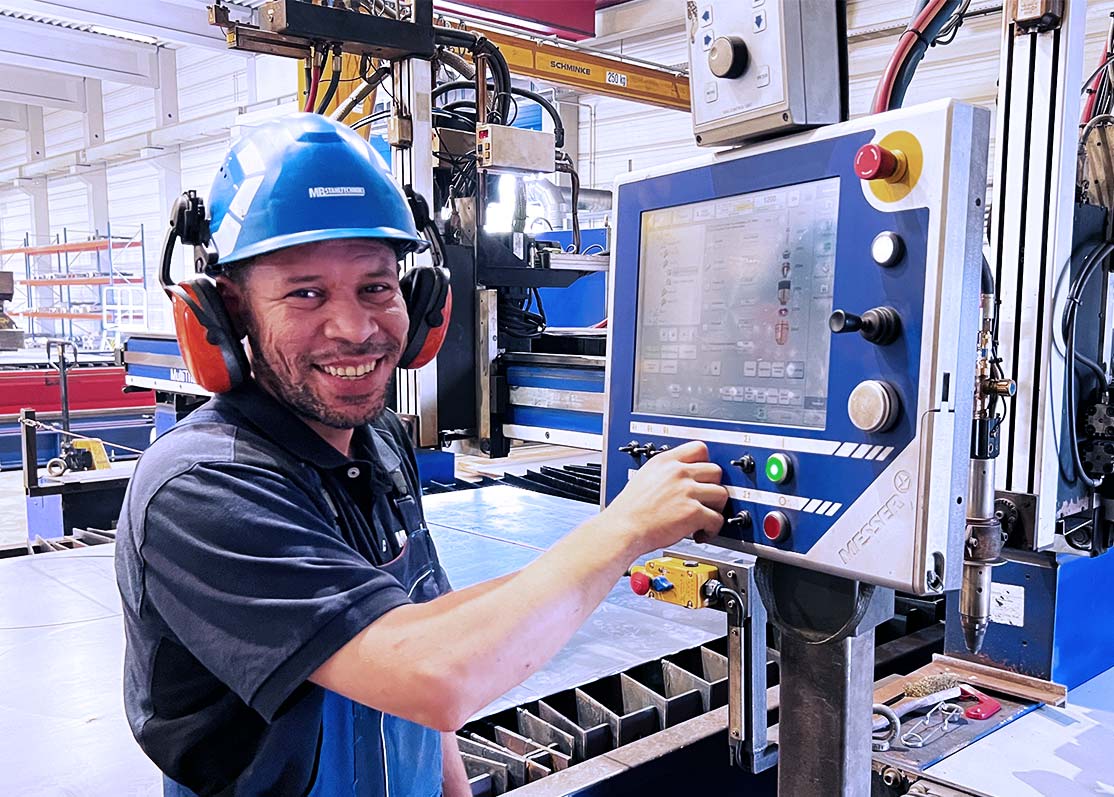Arbeiter mit Schutzhelm bedient ein großes Industriesteuerpult mit Touchscreen und Not‑Aus‑Taste.