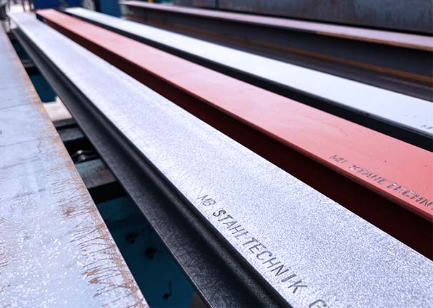 Mehrere parallel liegende Stahlträger, darunter ein rot grundierter Balken mit sichtbarer Beschriftung.
