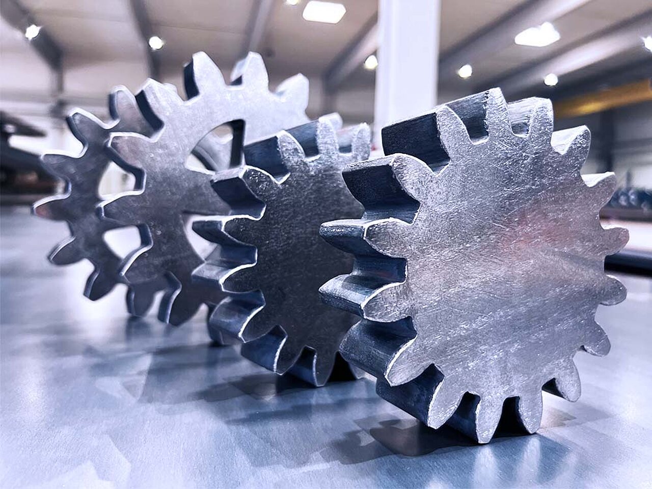 Drei polierte Metallzahnräder liegen auf einer reflektierenden Fläche in einer Industrieumgebung.