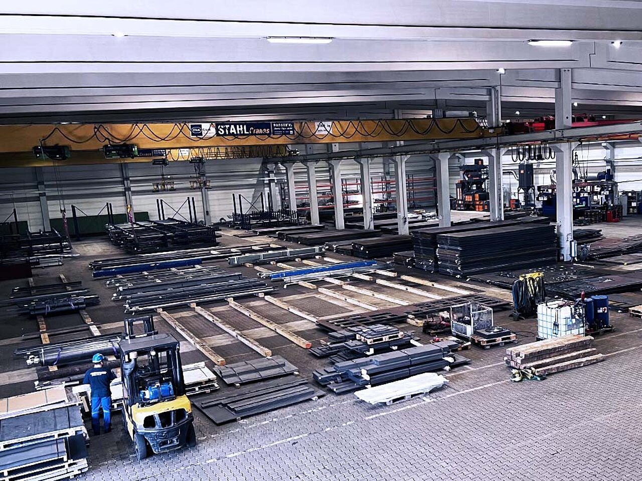 Große Industriehalle mit gestapelten Metallteilen, zwei Arbeitern und einem Gabelstapler unter einem STAHL‑Kran.